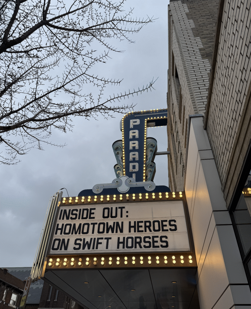 A theatre marquee reads: “INSIDE OUT: HOMOTOWN HEROES ON SWIFT HORSES” with the word “Paradise” in neon above. Bare tree branches and a cloudy sky are visible in the background.